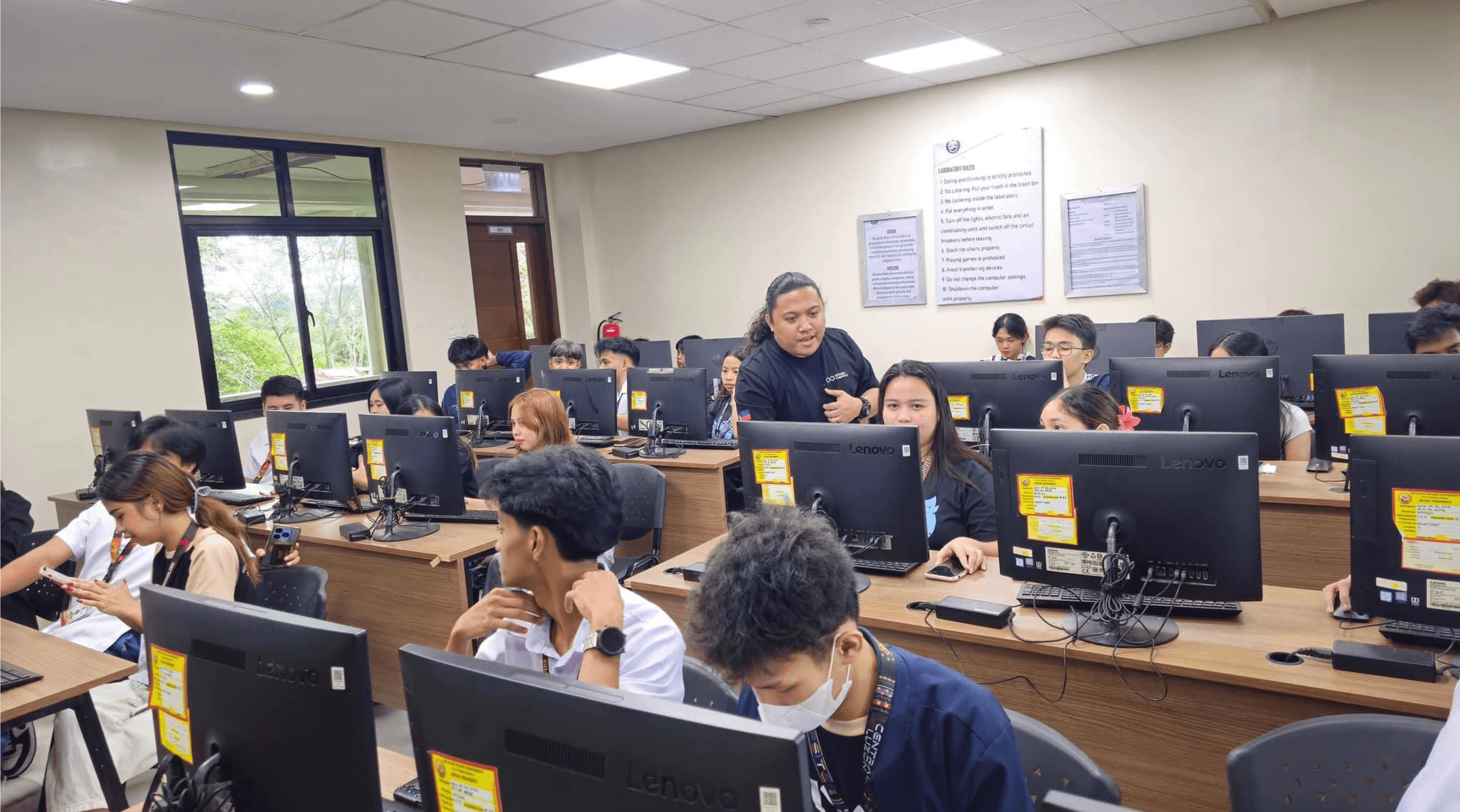 A DvCode facilitator leading a hands-on session in a university computer laboratory full of students at desktop machines.
